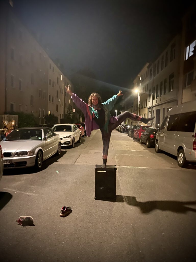 Josefine steht nachts lachend mit einem Bein hoch in der Luft auf ihrer Soundboks, die mitten auf der Straße steht