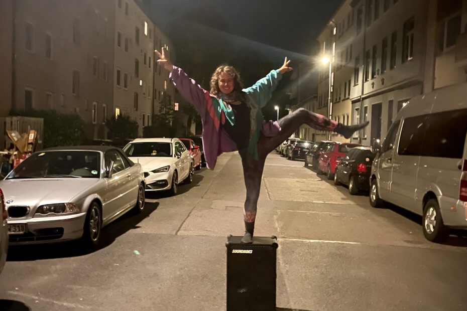 Josefine steht nachts mitten auf der Straße mit einem Bein hoch in der Luft auf ihrer Soundboks und feiert das Leben