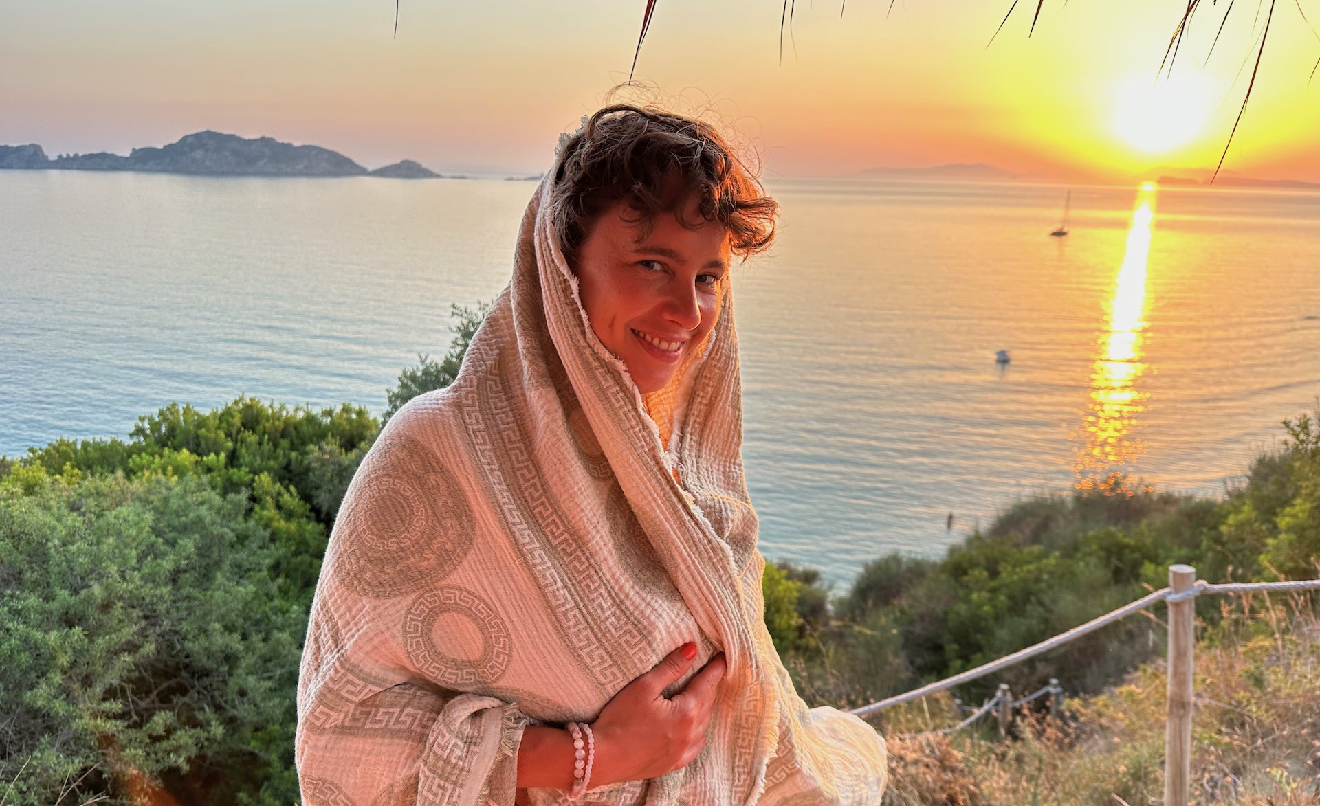 Porträt von Josefine Puppe mit einem Tuch halb über den Kopf gedeckt, lachend vor Sonnenuntergang am Meer, einladender Blick