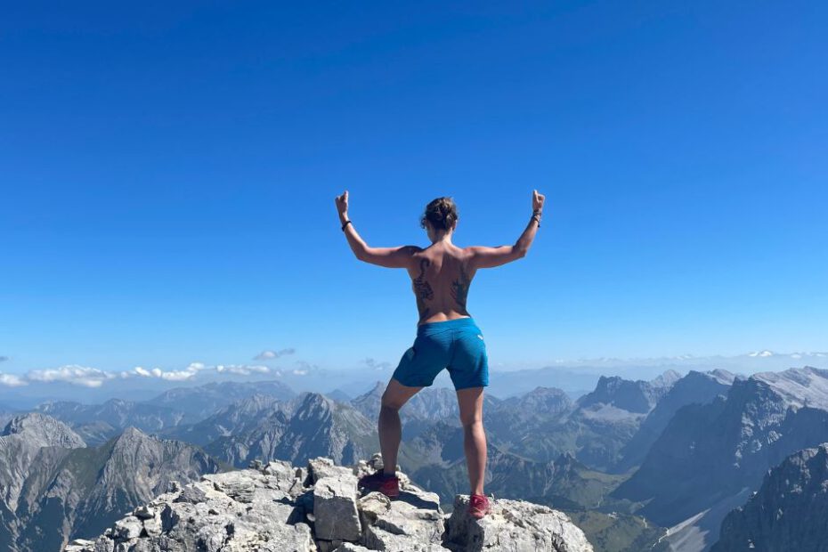 Josefine Puppe steht mit erhobenen Armen auf einem Berggipfel – Symbol für Freiheit, Stärke und das Überwinden von Scham.