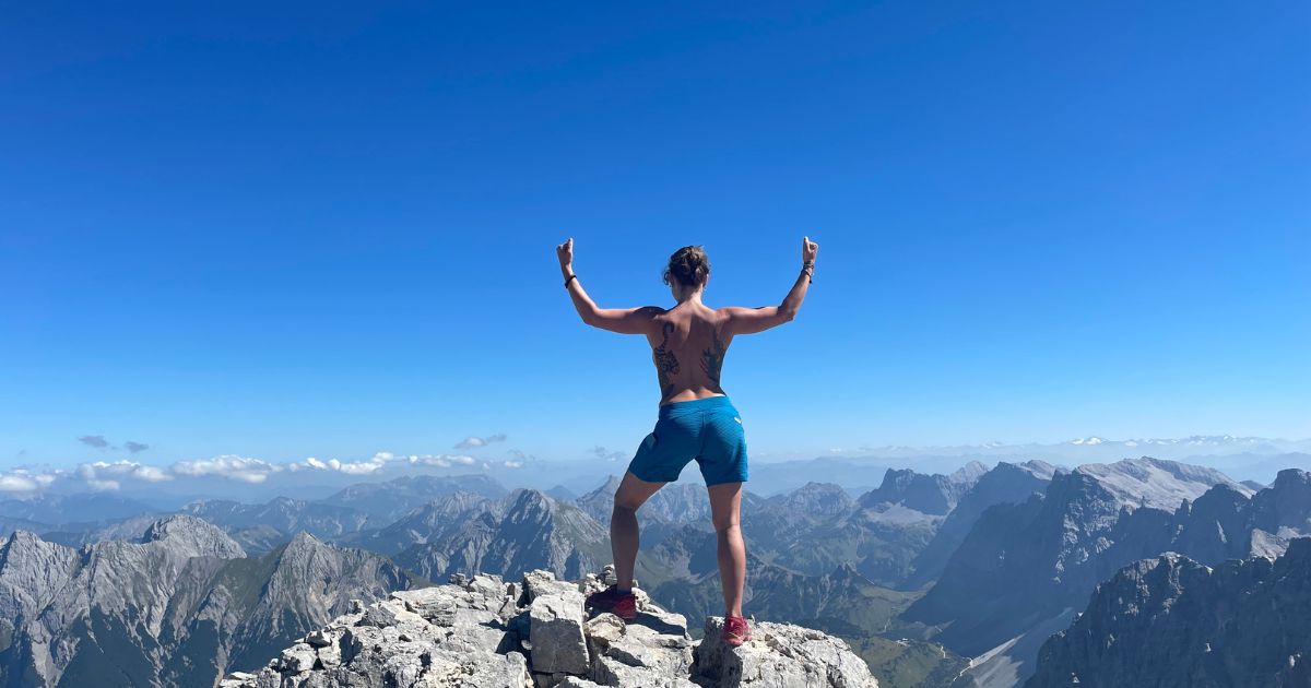 Josefine Puppe steht mit erhobenen Armen auf einem Berggipfel – Symbol für Freiheit, Stärke und das Überwinden von Scham.